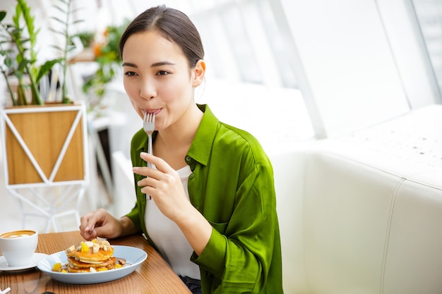 Smiling asian woman having pancakes for breakfast at the cafe indoors