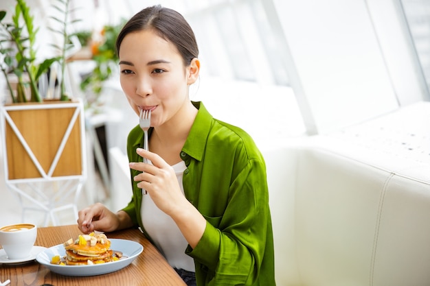 Sorridente donna asiatica che mangia frittelle per colazione al bar al chiuso