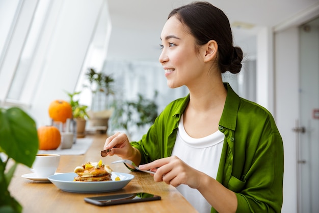 Smiling asian woman having pancakes for breakfast at the cafe indoors