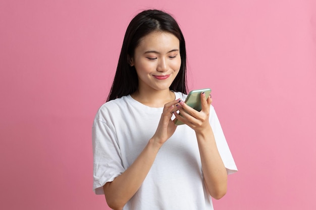 Smiling asian woman is pointing on smartphone standing on pink background