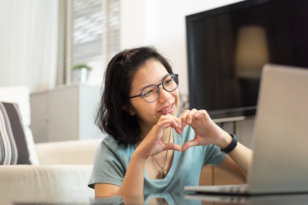 写真 ノートパソコンとのビデオ通話中にハートの形で彼女の手を作る笑顔のアジア女性