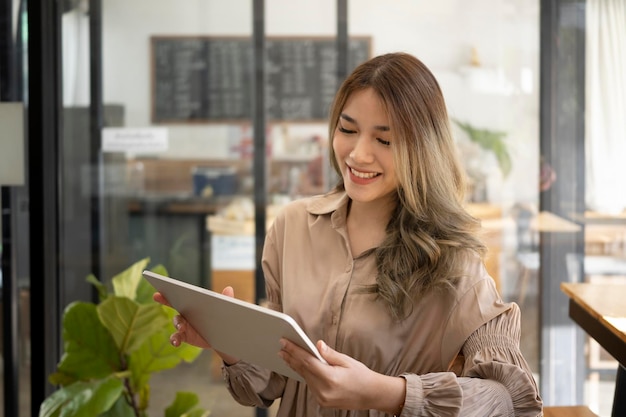 Smiling asian woman owner standing at her coffee shop and using digital tablet