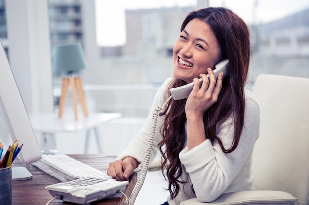 オフィスのコンピューターを使用して電話でアジアの女性の笑みを浮かべてください。