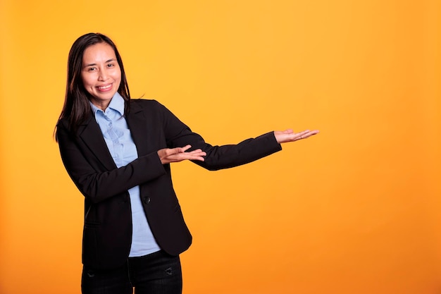 Smiling asian woman pointing to side with forefinger, advertising new product in studio over yellow background. adult in formal suit standing with positive facial expression, during advertisement job