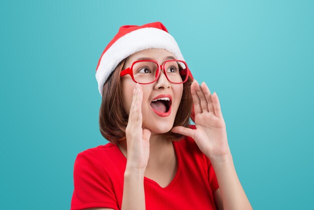 Smiling asian woman portrait with christmas santa hat shouting isolated on blue background.