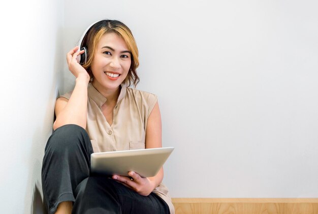 Smiling Asian woman portrait with short hair in beige sleeveless shirt relax with music or watching movie on tablet and wearing white wireless headphone on white wall background with copy space