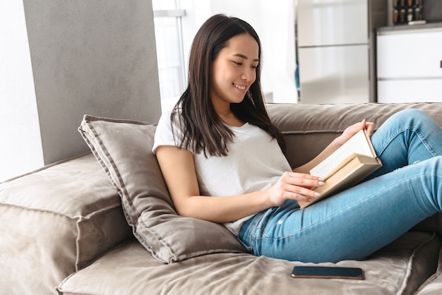 Sorridente donna asiatica che legge un libro a casa, rilassandosi su un divano