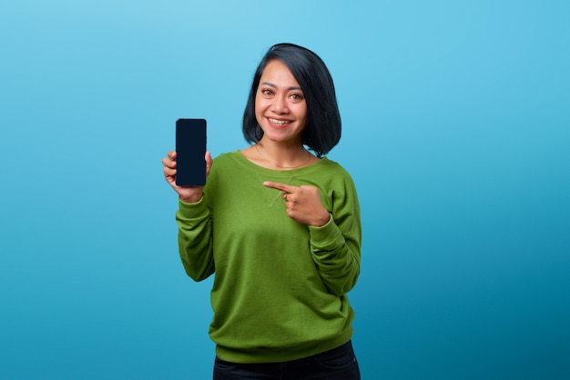 Smiling asian woman showing blank smartphone screen and pointing on it while looking at the camera