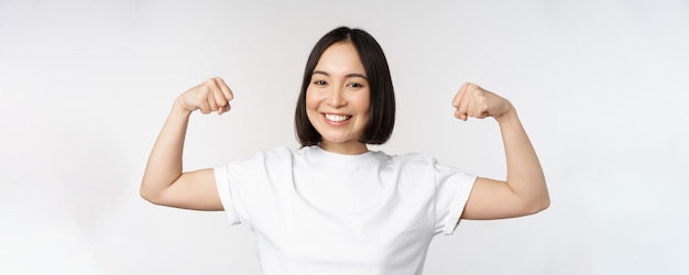 Smiling asian woman showing flexing biceps muscles strong arms gesture standing in white tshirt over white background