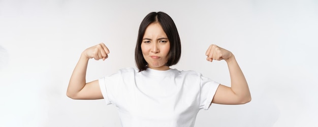 Улыбающаяся азиатка показывает сгибание мышц бицепса сильным жестом рук, стоя в белой футболке над