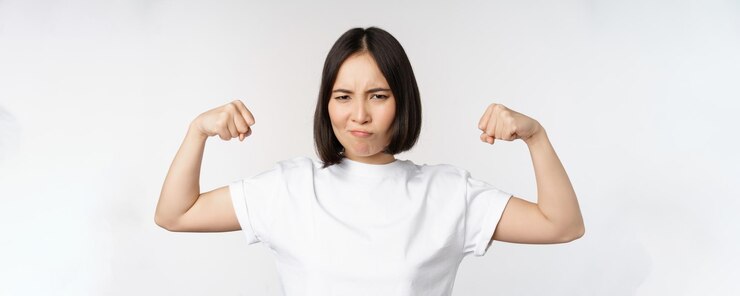 Photo 上腕二頭筋の屈曲を示す笑顔のアジアの女性は、白いtシャツの上に立っている強い腕のジェスチャー