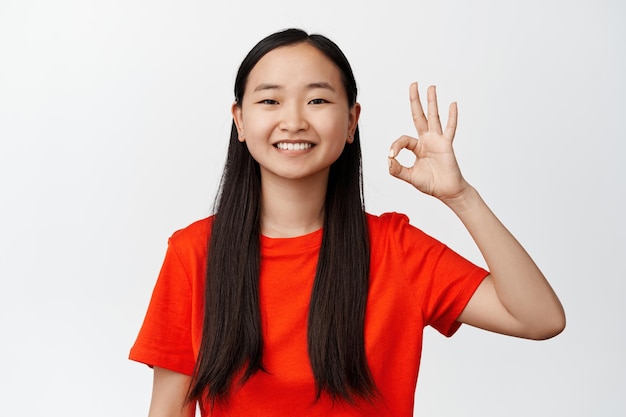 Smiling asian woman showing okay ok sign and looking satisfied recommending good quality standing against white background