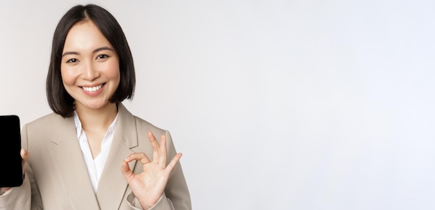 Photo smiling asian woman showing smartphone screen and okay sign corporate person demonstrates mobile phone app interface standing over white background