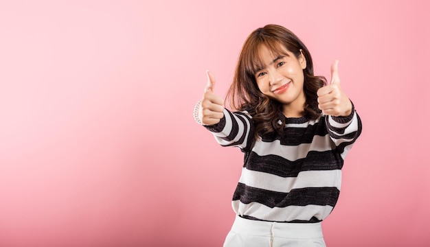 A smiling Asian woman in a studio portrait on pink background makes a thumbsup 'Ok' sign symbolizing agreement and success Her cheerful expression radiates positivity and satisfaction