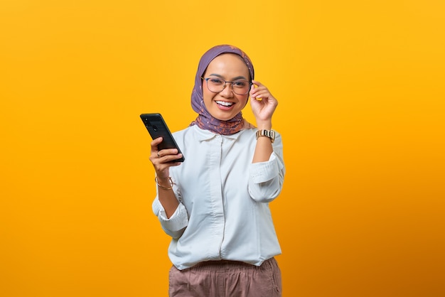 Smiling Asian woman touching glasses and holding mobile phone in one hand over yellow background