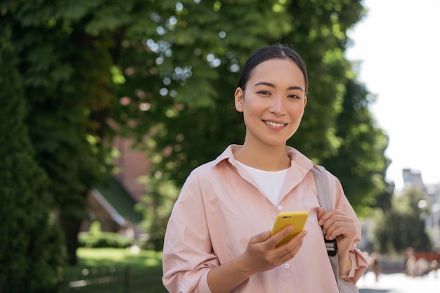 Smiling asian woman using mobile phone looking at camera walking in park