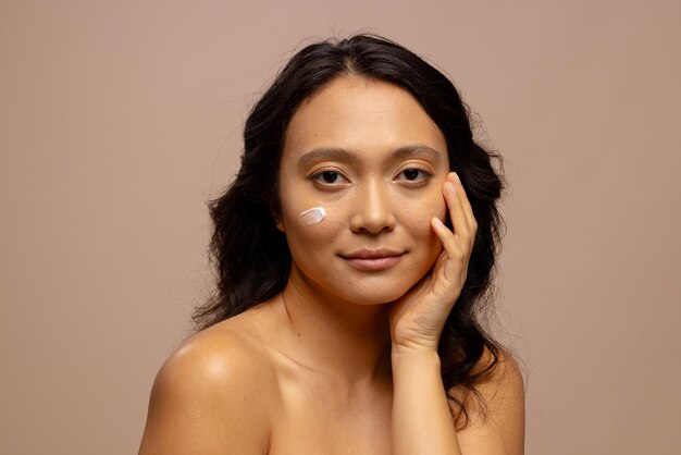 Smiling asian woman with dark hair with skin cream on her cheek touching face with hand