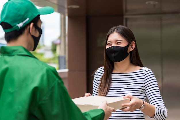 Smiling asian woman with face mask take pizza from deliveryman\
in green jacket uniform. prevent covid-19 delta pandemic. new\
normal to eat from home and order food online by phone app.