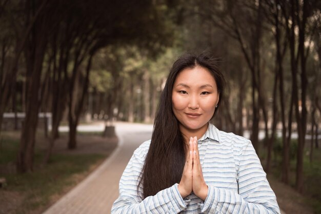 Smiling asian woman with hands in namaste jesture in city park at summer