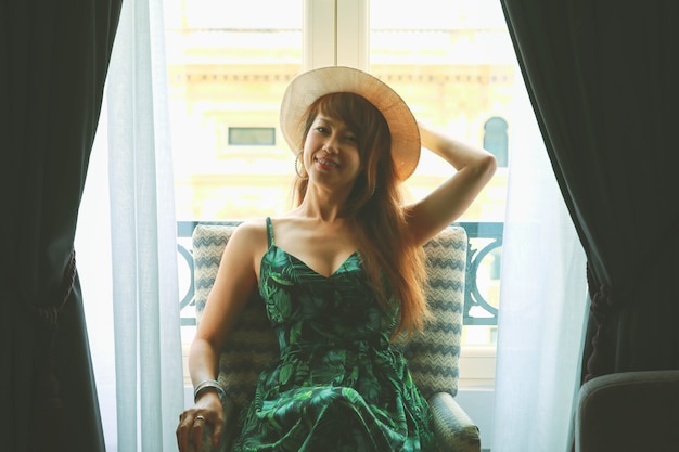 Smiling asian woman with hat siting on chair, feeling happy and positive