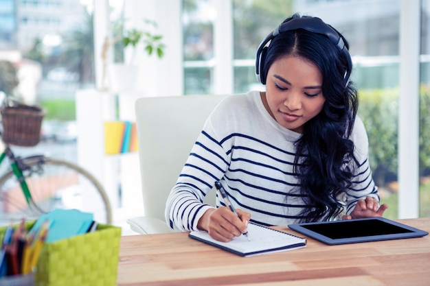 Smiling Asian woman with headphones writing on notepad in office