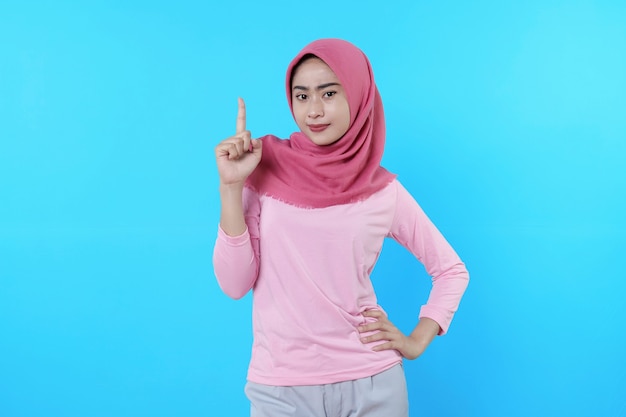 Smiling asian woman with her finger pointing isolated on light blue background wearing hijab with pink t shirt