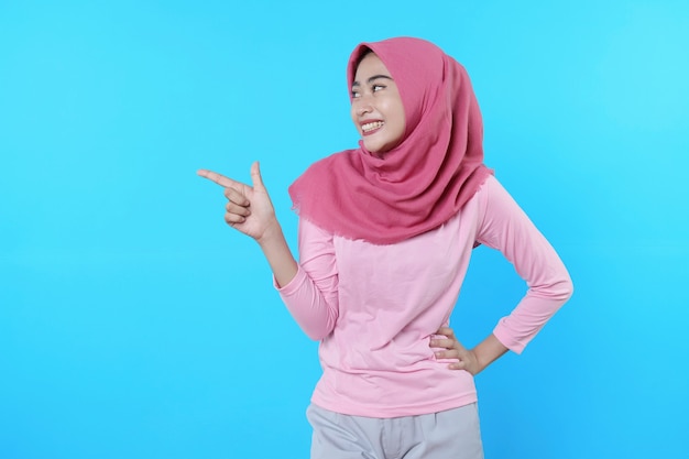 Smiling asian woman with her finger pointing isolated on light blue background wearing hijab with pink t shirt