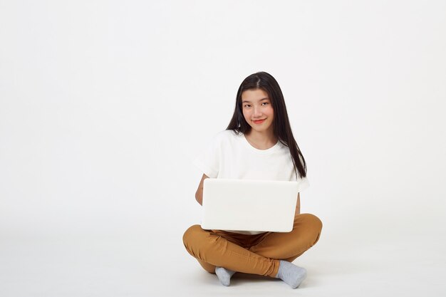 Smiling Asian woman working on laptop computer while sitting on the floor with legs crossed isolated over white âbackground, Back to school concept