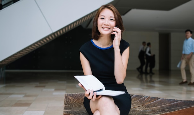 검은 정장을 입은 아시아 젊은 여성 사업가가 무릎을 꿇고 다리를 꼬고 있습니다.