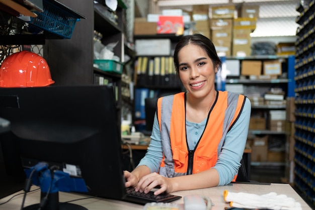 Foto sorridente giovane donna asiatica seduta e utilizzando il computer desktop in fabbrica magazzino