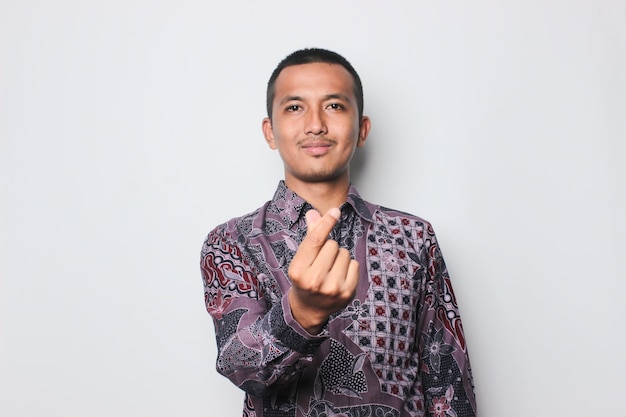 Улыбающийся азиатский молодой человек в рубашке из батика с символом руки любви на белом фоне