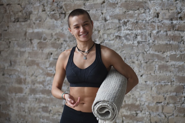 Photo smiling athletic woman with yoga exercise mat standing near brick wall, looking at camera