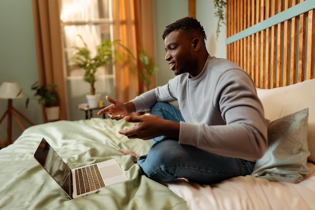 Smiling attractive African American man sitting on bed at home using laptop having video conference