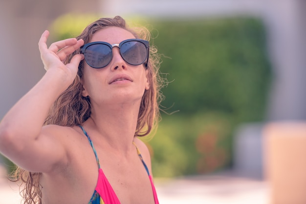 緑の木の背景にサングラスをかけた魅力的なブロンドの巻き毛の女性を笑顔。