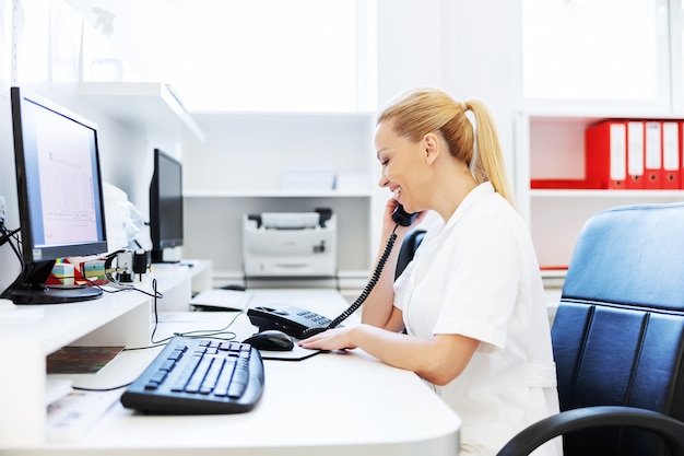 Smiling attractive blond female lab assistant in white sterile uniform sitting in laboratory and having phone call from patient.