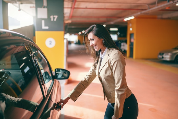 Smiling attractive brunette dressed smart casual opening car door at public garage.