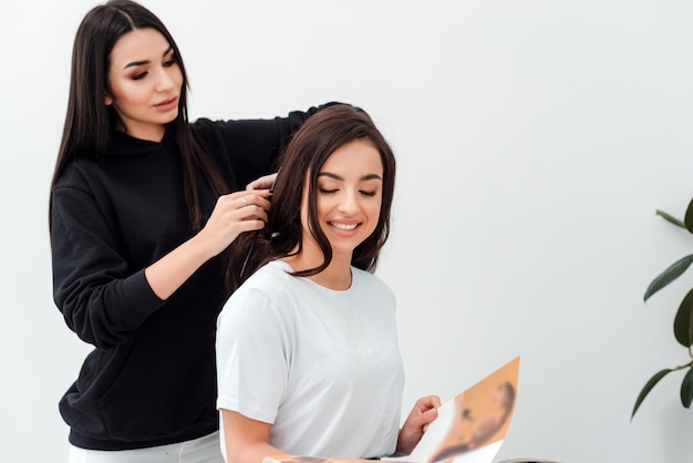 Smiling, attractive brunette is reading a magazine while her hairdresser combs her beautiful hair