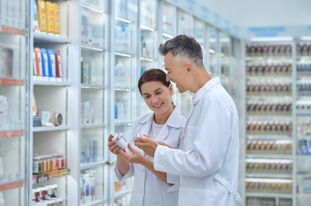 Smiling attractive female druggist and her cheerful male colleague holding medicament bottles in their hands