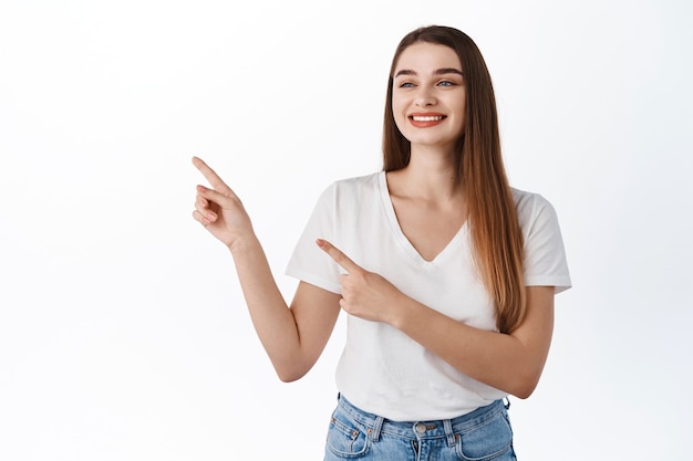 Sorridente ragazza attraente che indica, guardando da parte con faccia compiaciuta, guardando l'offerta promozionale, mostrando pubblicità sullo spazio della copia, in piedi in maglietta contro il muro bianco