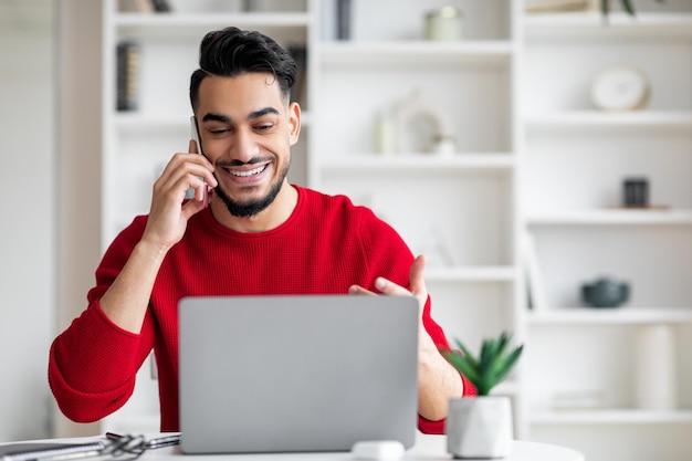 Smiling attractive millennial arab male with beard in red clothes speaks by phone and looks at