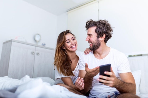 一緒にベッドに横たわっているスマートフォンを使用して魅力的な千年紀のカップルを笑顔、朝にソーシャルネットワークのニュースをチェックしている幸せなボーイフレンドとガールフレンド、男性に新しい携帯電話アプリを見せている若い女性