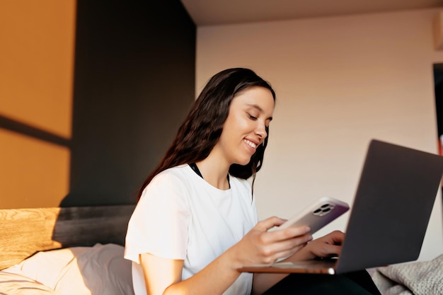 Smiling attractive pretty woman with happy smile in white shirt is sitting on the bed in sunlight and typing on laptop Girl is holding zoom conference on laptop with phone