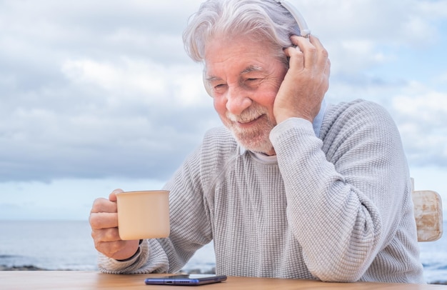 Uomo in pensione adulto senior attraente sorridente con la barba che tiene una tazza di caffè all'aperto in mare