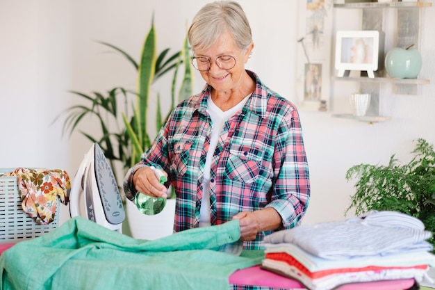 Smiling attractive senior woman in checkered shirt and glasses ironing clothes at home on ironing board