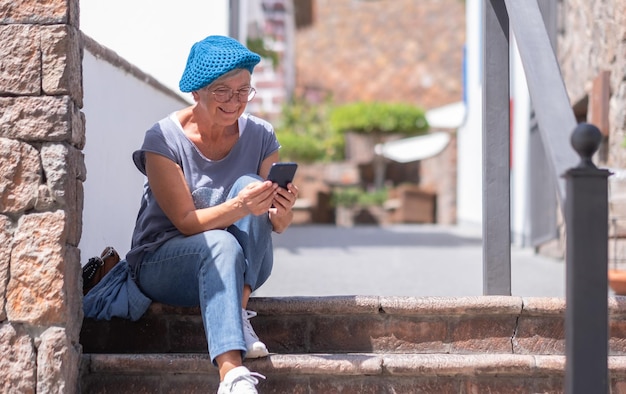 Smiling attractive senior woman with blue cap sitting outdoors on staircases using mobile phone Elderly lady relaxing enjoying vacation or retirement under the sun