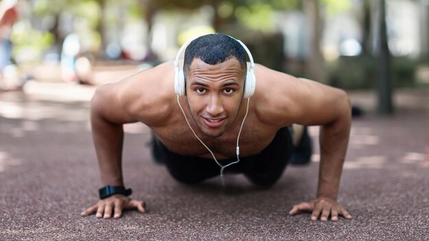 Фото Улыбающийся привлекательный сильный черный парень в наушниках слушает музыку, делая отжимания в парке по утрам