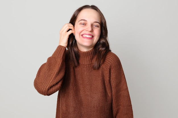 Smiling attractive winsome Caucasian woman wearing brown sweater standing isolated over light gray background looking at camera with happy expression gently touching her hair