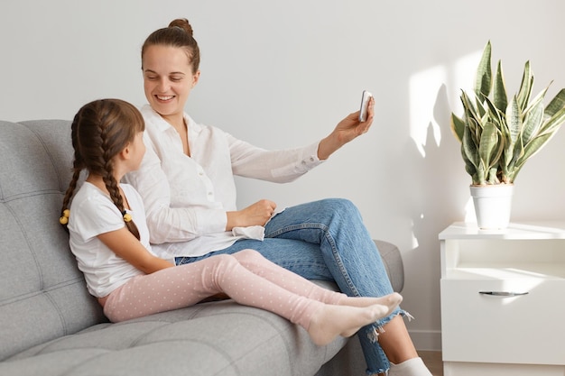 Smiling attractive young adult female wearing white shirt sitting on sofa with her daughter and holding smartphone in hands, mommy asking her child to make selfie.