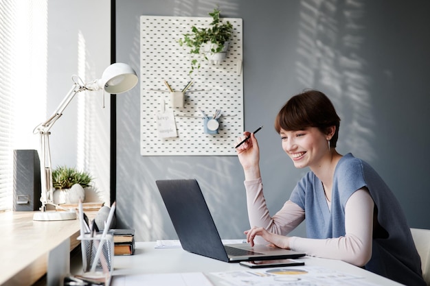 Smiling attractive young businesswoman with short hair sitting in home office and using laptop while