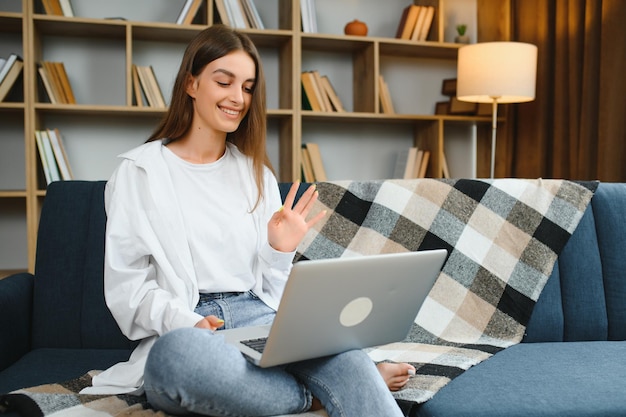 Smiling attractive young woman sitting on sofa using laptop\
communicating working online at home happy teen girl typing on\
computer enjoying writing blog or chatting with friends in social\
network